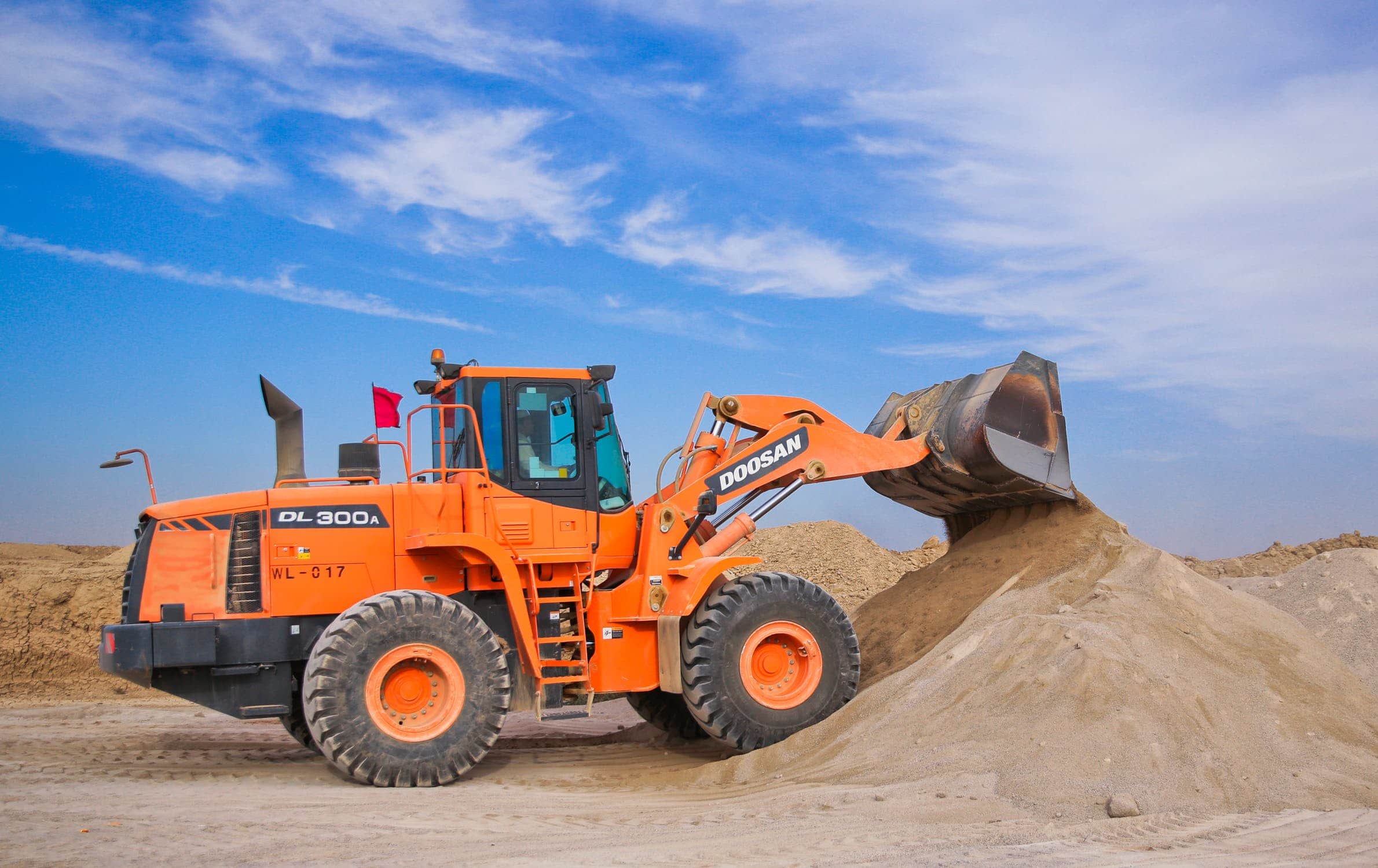 Orange Doosan DL300A wheel loader scooping sand on a construction site under a blue sky.