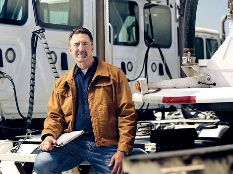 Man in brown jacket holding clipboard, seated near white fleet trucks and tanker trailer